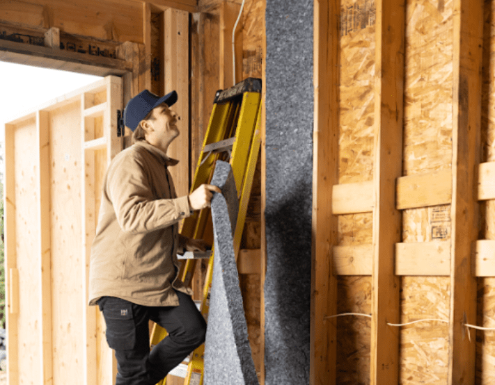 Worker installing denim insulation batts between wooden wall studs.